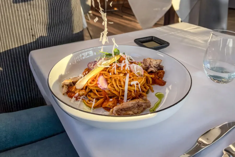 Ein Kellner streut frischen Parmesan über einen tiefen Teller mit Pasta und Fleisch an einem elegant gedeckten Tisch.