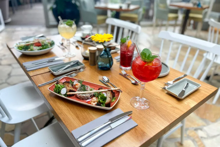 Draufsicht auf zwei hölzerne Restauranttische mit bunten Tellern, Cocktails, Besteck, einer gelben Blume und einer Kerze.