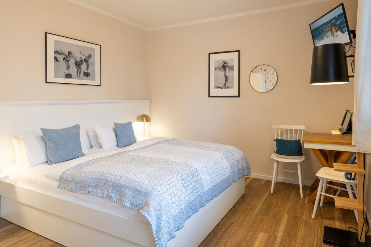 Doppelzimmer Petit im STRANDHAUS Ahrenshoop mit Doppelbett, Schreibtisch, weißem Stuhl und Strandfotografien.