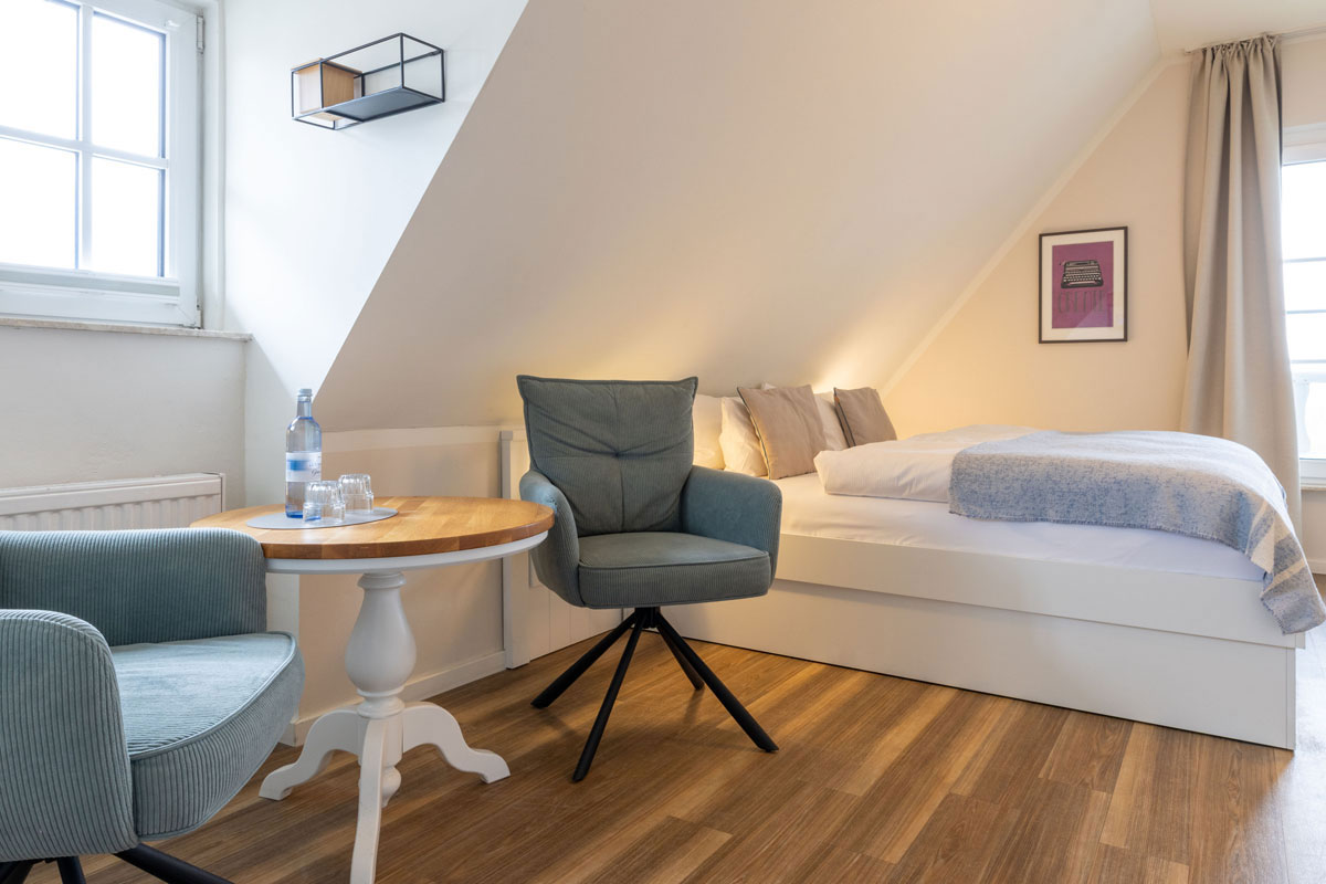 Sitzecke im Doppelzimmer Petit des STRANDHAUS Ahrenshoop mit zwei Sesseln, rundem Tisch und Doppelbett unter der Dachschräge.
