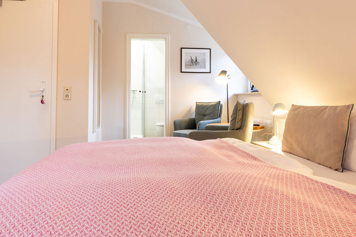 Doppelzimmer Petit im STRANDHAUS Ahrenshoop mit rosafarbener Decke, Sessel, Stehlampe und Blick ins Bad.
