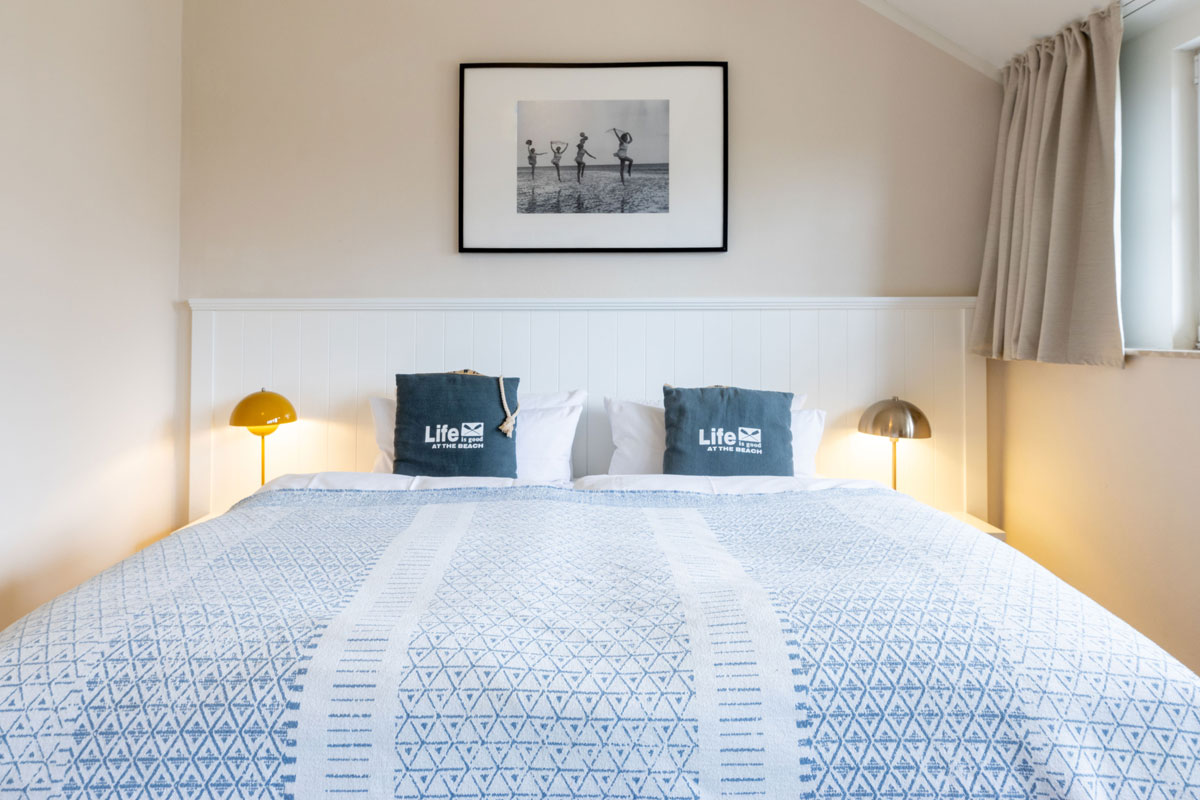 Doppelbett im STRANDHAUS Ahrenshoop mit blauen Kissen, warmem Licht und Strandfotografie an der Wand.
