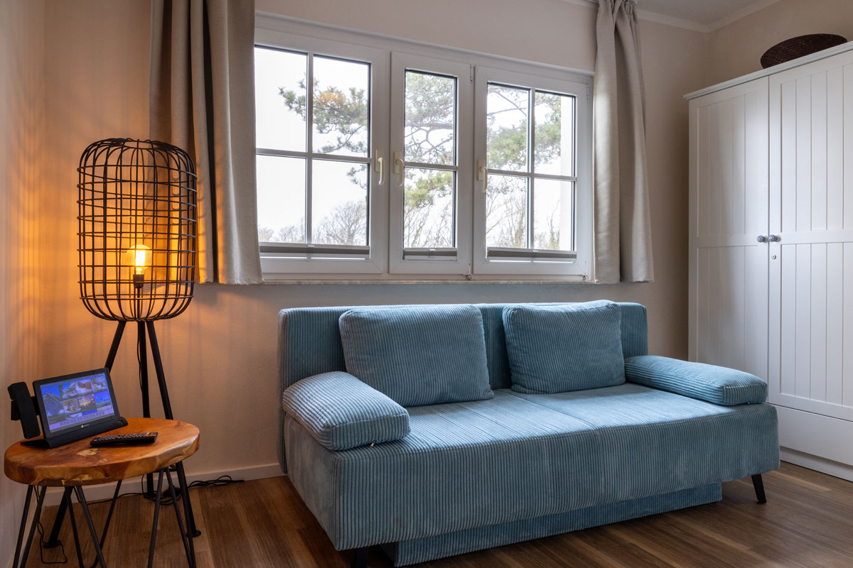Sitzbereich im Doppelzimmer des STRANDHAUS Ahrenshoop mit blauem Sofa, Stehlampe und weißem Kleiderschrank.