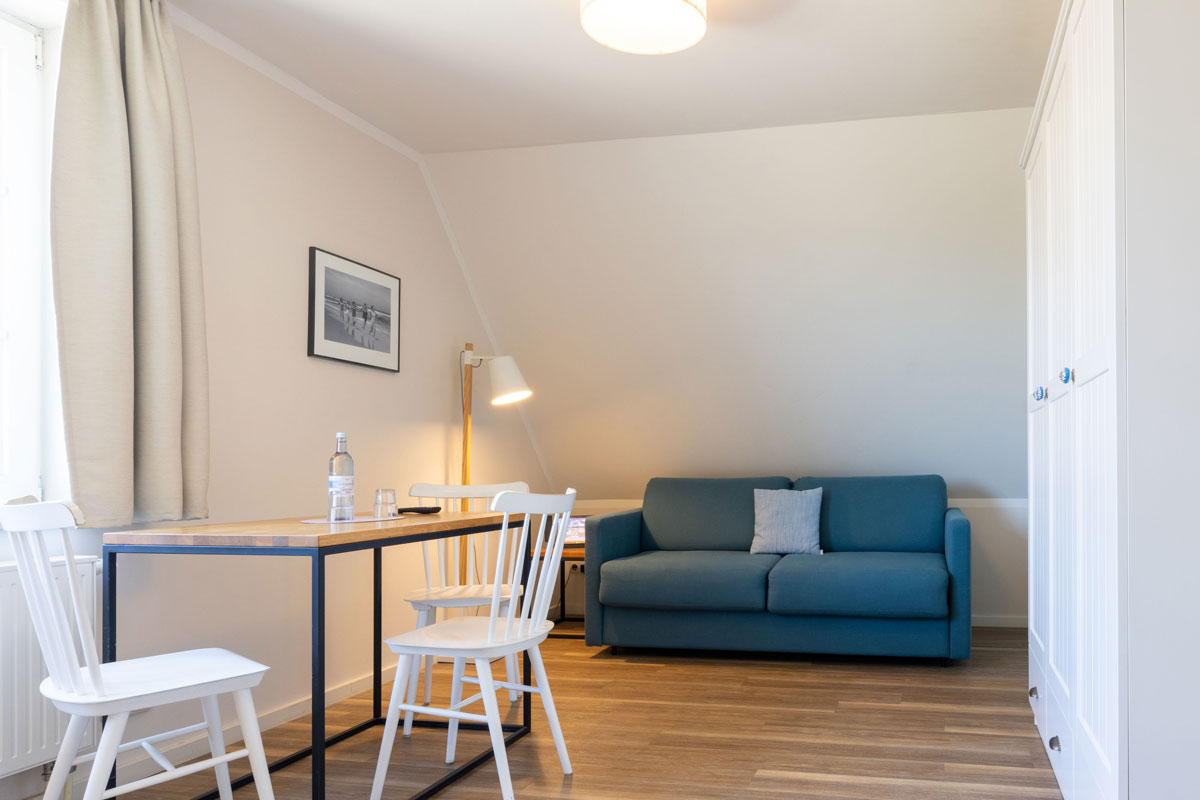 Wohnbereich im Doppelzimmer des STRANDHAUS Ahrenshoop mit Esstisch, weißen Stühlen und blauem Sofa.