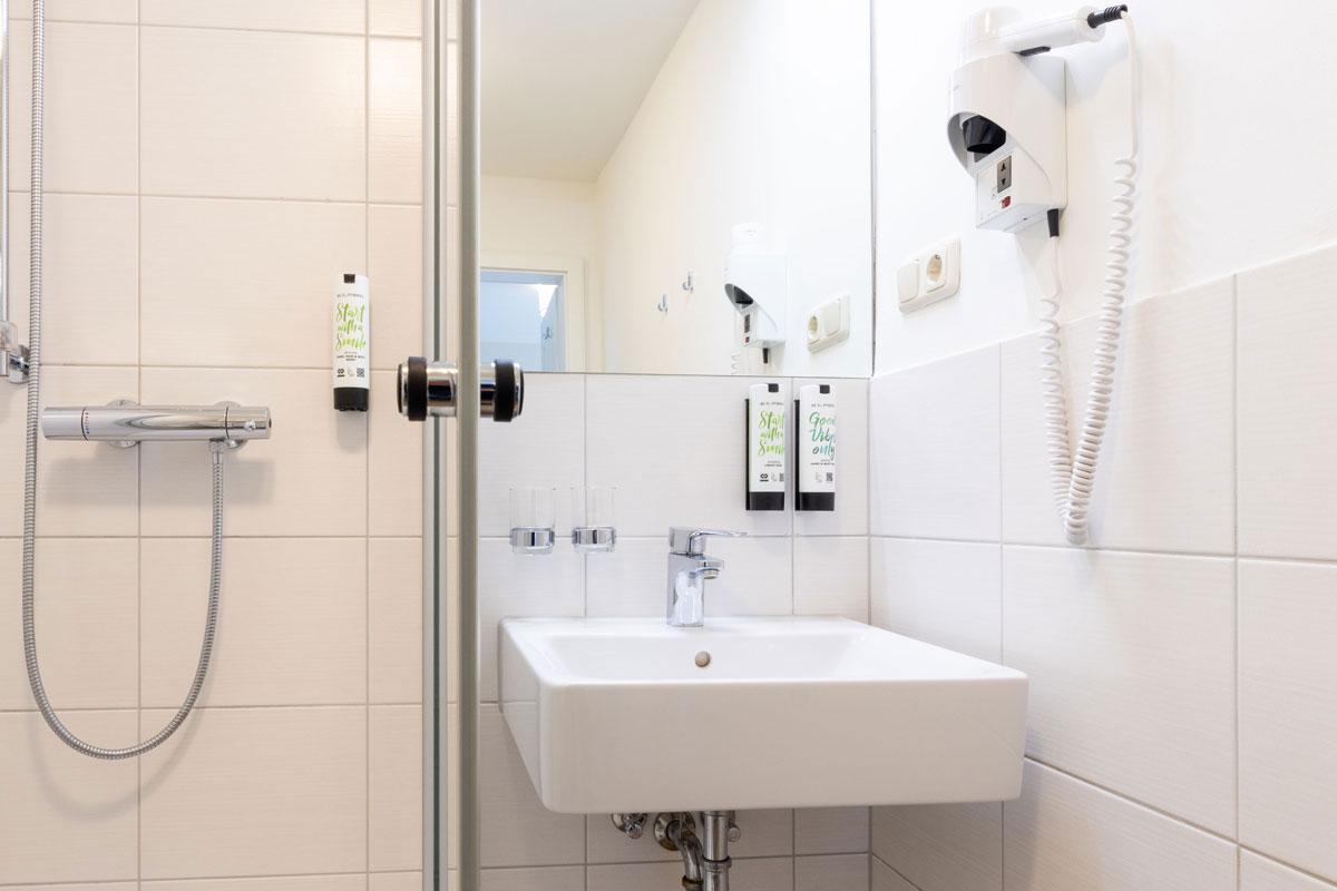 Bad im Einzelzimmer des STRANDHAUS Ahrenshoop mit Glasdusche, Waschbecken, Spiegel und Haartrockner.