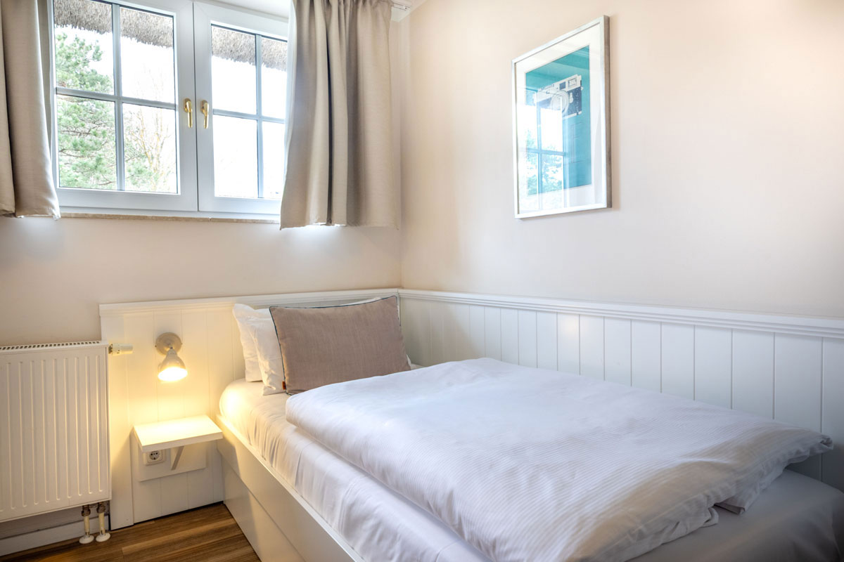 Einzelbett im STRANDHAUS Ahrenshoop mit warmer Wandlampe, weißer Holzverkleidung und Fenster zum Grünen.