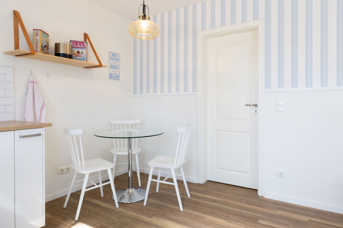 Essbereich im Apartment des STRANDHAUS Ahrenshoop mit rundem Glastisch, weißen Stühlen und blau-weißer Streifentapete.