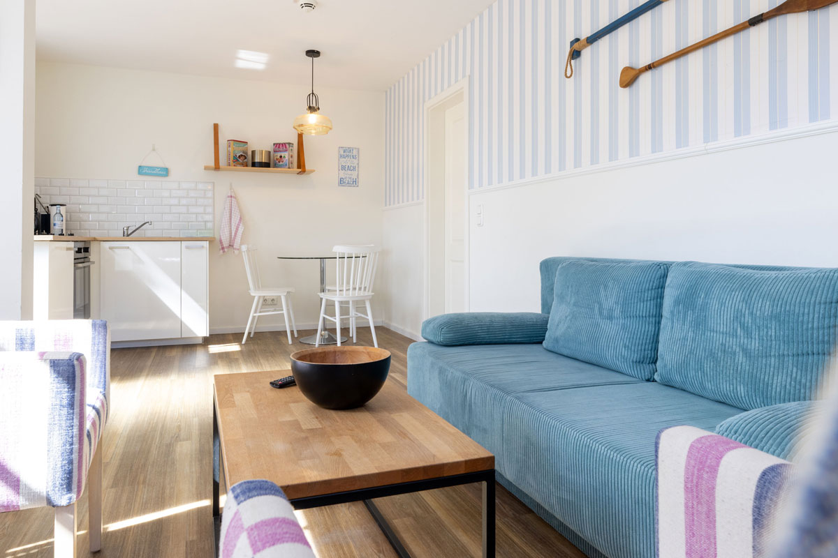 Wohnbereich im Apartment des STRANDHAUS Ahrenshoop mit blauem Sofa, Couchtisch, Esstisch und offener Küche.