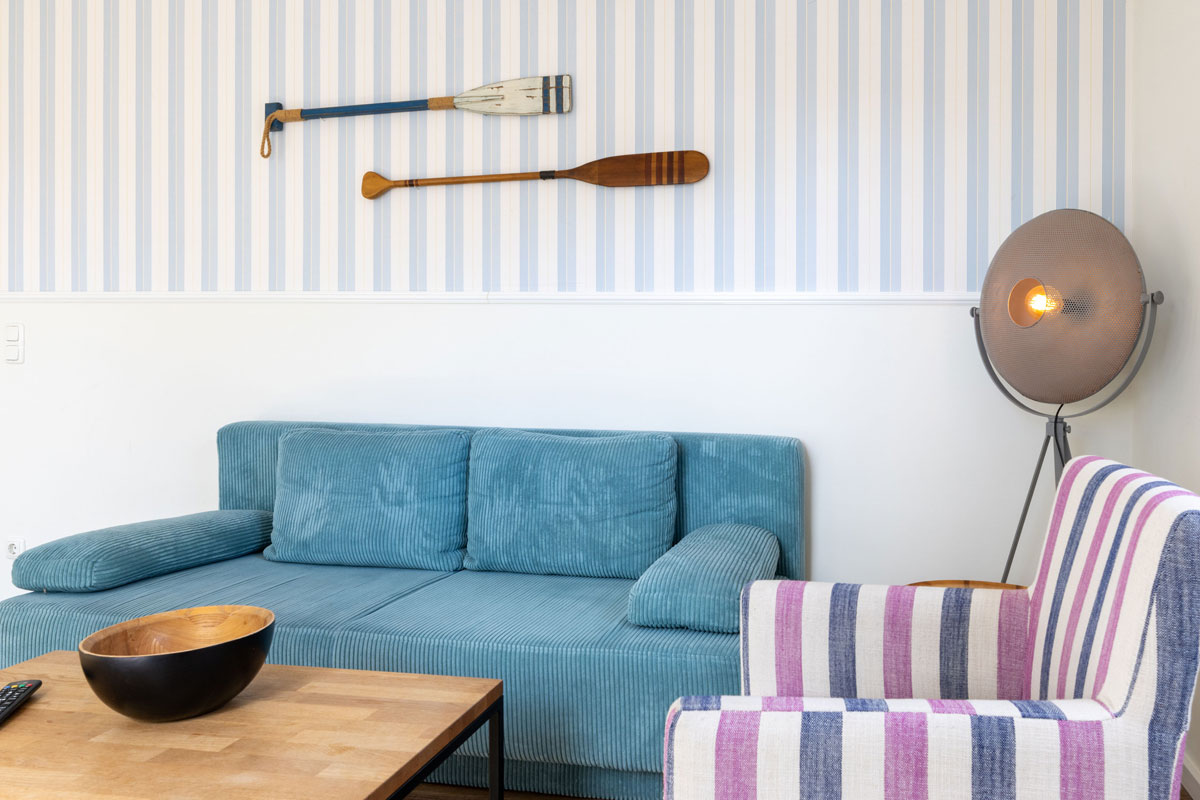 Sitzecke im Apartment des STRANDHAUS Ahrenshoop mit blauem Sofa, gestreiftem Sessel und Holzrudern an der Wand.