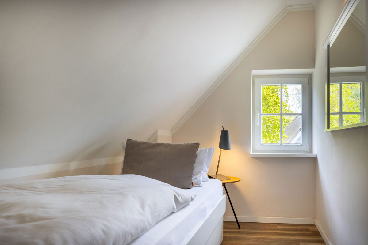 Einzelbett im Familienzimmer Petit des STRANDHAUS Ahrenshoop mit Nachttischlampe, Dachschräge und Fenster zum Grünen.