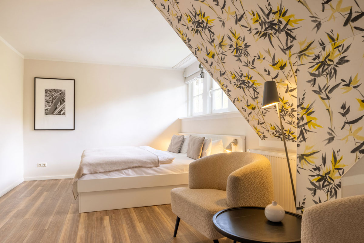 Gesamtansicht des Familienzimmers Petit im STRANDHAUS Ahrenshoop mit Doppelbett, Sesseln, Stehlampe und floralem Tapetenmotiv.