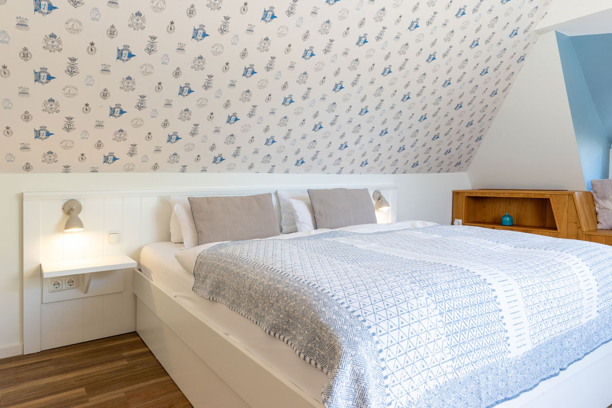 Doppelbett im Dachzimmer Landseite des STRANDHAUS Ahrenshoop mit maritimer Tapete, Wandlampen und Holzeinbauten.
