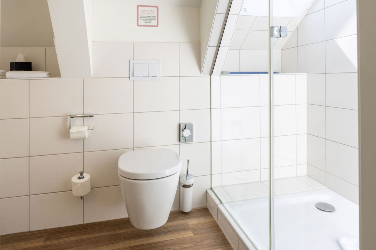 Bad im Dachzimmer Meerseite des STRANDHAUS Ahrenshoop mit Glasdusche, WC und weißen Fliesen unter der Dachschräge.