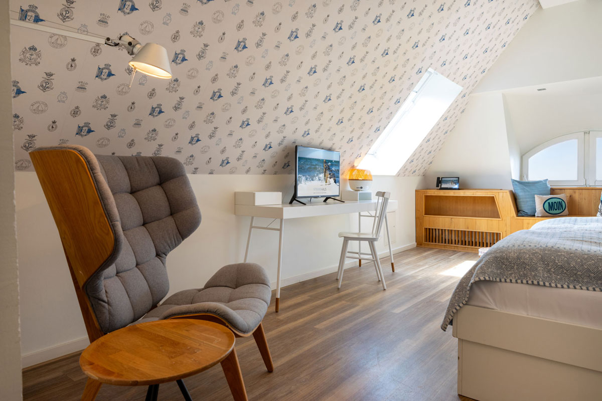 Dachzimmer Meerseite im STRANDHAUS Ahrenshoop mit grauem Sessel, Schreibtisch, Bett und Holzbank mit Meerblick.