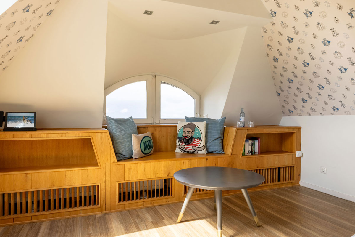 Holzbank mit Meerblick durch das Rundbogenfenster im Dachzimmer Meerseite des STRANDHAUS Ahrenshoop mit maritimen Kissen und Couchtisch.