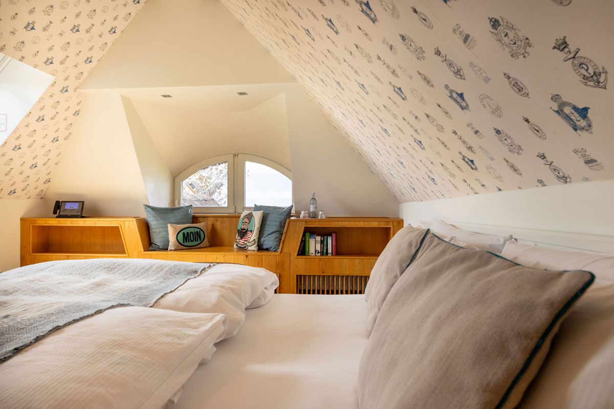 Schlafbereich im Dachzimmer Meerseite des STRANDHAUS Ahrenshoop mit Doppelbett, Holzbank und Rundbogenfenster mit Meerblick.