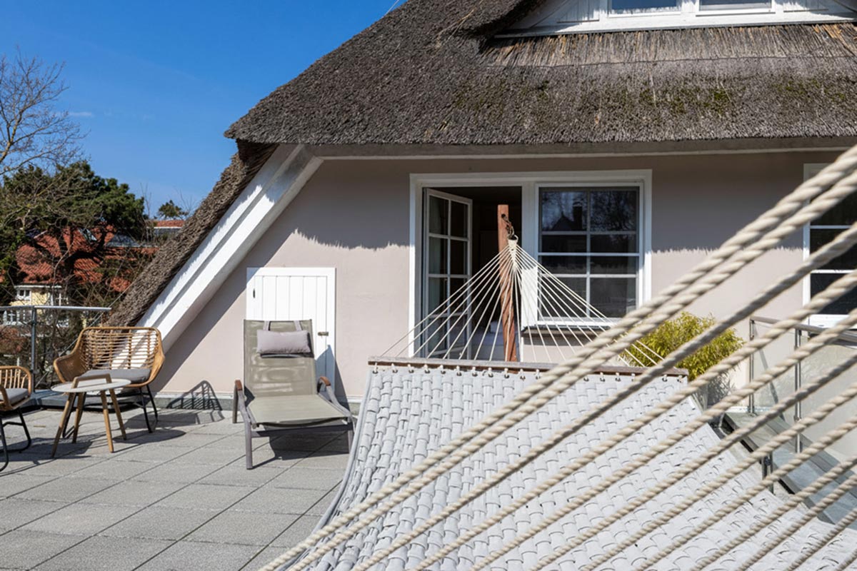 Sonnendeck des STRANDHAUS Ahrenshoop mit Hängematte, Liegestuhl, Rattansessel und Reetdach im Hintergrund.