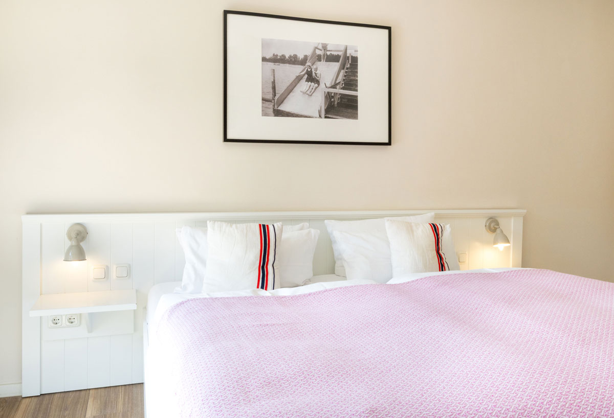 Doppelbett im Doppelzimmer Sonnendeck des STRANDHAUS Ahrenshoop mit rosafarbener Decke, Wandlampen und schwarz-weißer Fotografie.