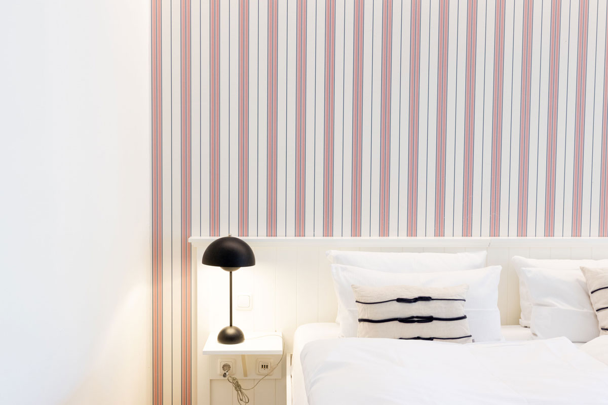 Detailaufnahme des Schlafbereichs der Terrassensuite im STRANDHAUS Ahrenshoop mit weißem Bett, Nachttischlampe und Streifentapete.