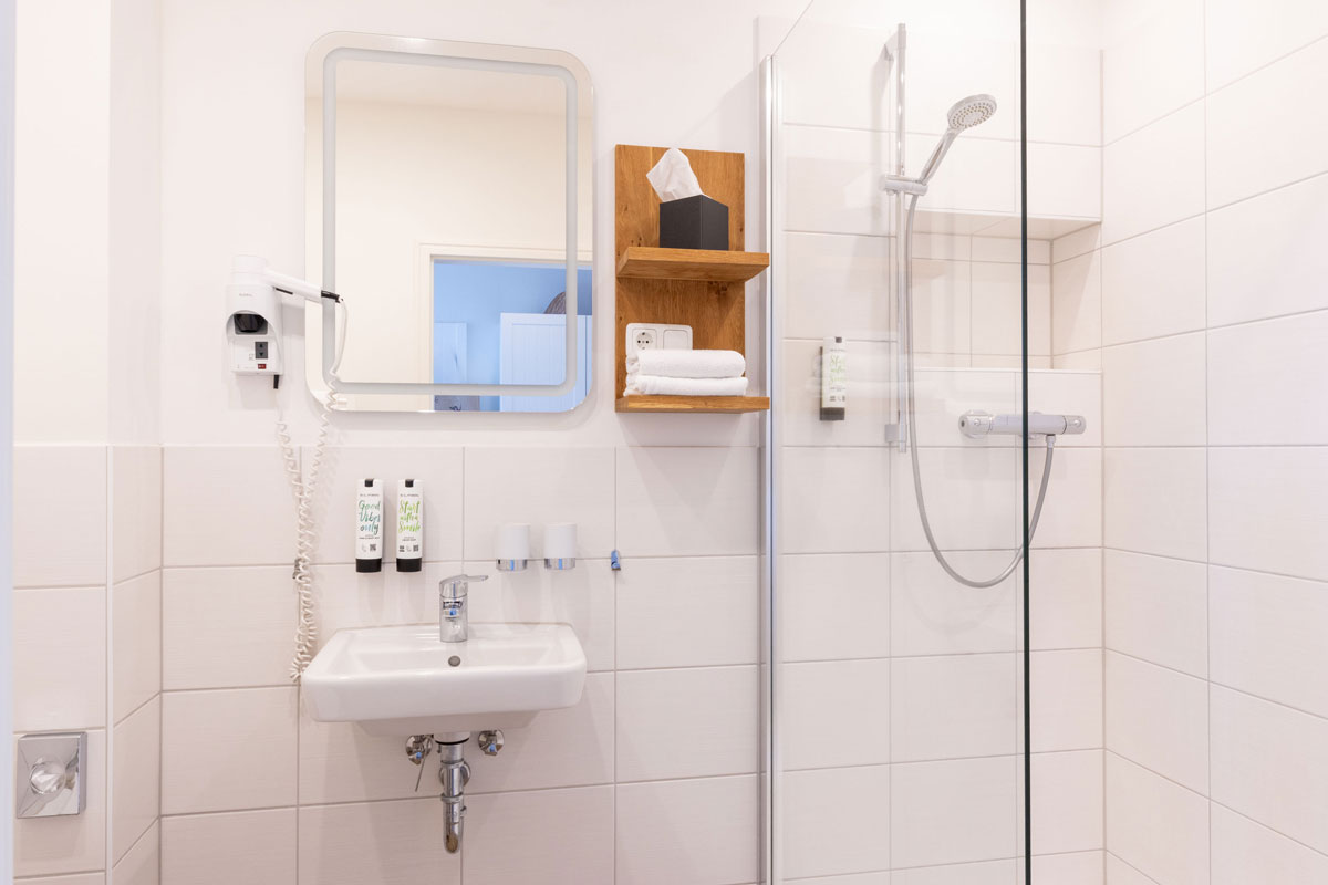 Badezimmer der Terrassensuite im STRANDHAUS Ahrenshoop mit Dusche, Waschbecken, Spiegel und Holzregal mit Handtüchern.