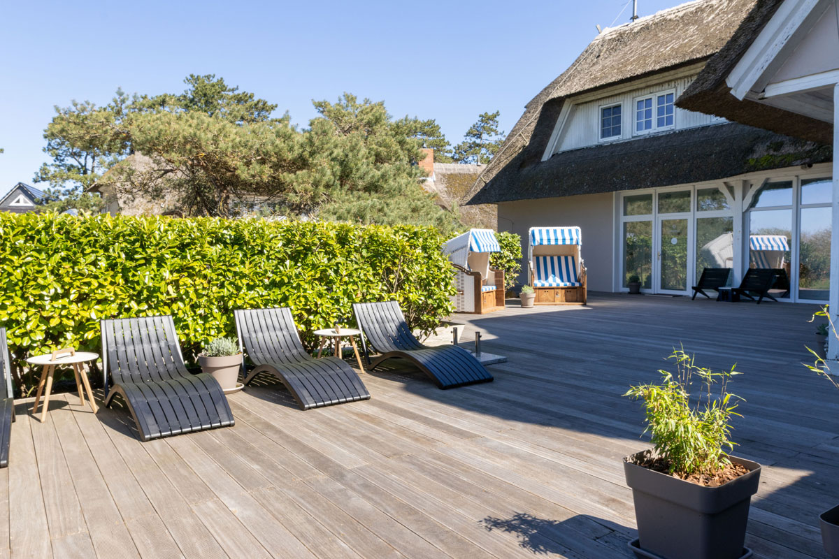 Sonnenterrasse des STRANDHAUS Ahrenshoop mit Liegestühlen, Strandkorb und Reetdach bei blauem Himmel.