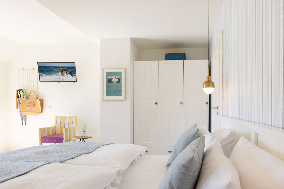 Terrassenzimmer im STRANDHAUS Ahrenshoop mit Doppelbett, weißem Kleiderschrank, gestreiftem Sessel und Wandfernseher.