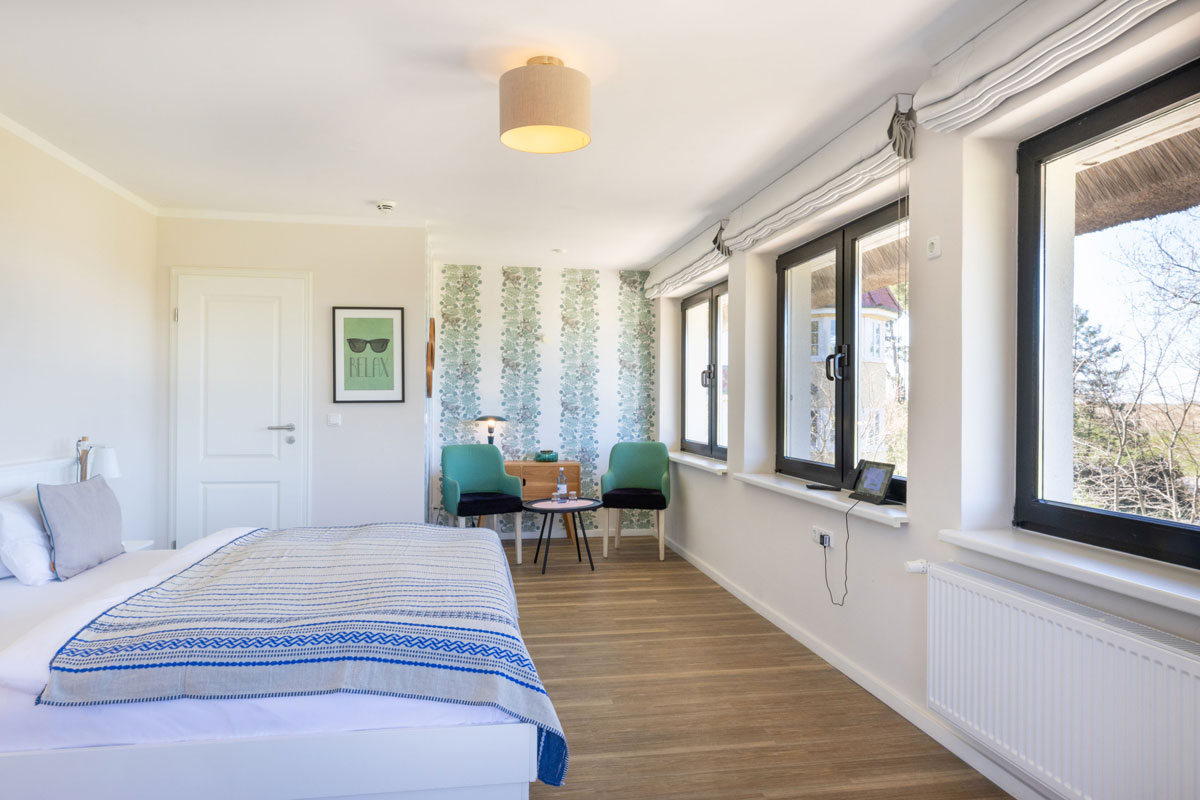 Terrassenzimmer im STRANDHAUS Ahrenshoop mit Doppelbett, Sitzecke mit grünen Sesseln und großen Fenstern mit Ausblick.